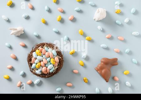 Easter composition of nest with pastel chocolate eggs, sweets, chocolate bunny and white ceramic bunny and on blue background. View from above. Flat l Stock Photo