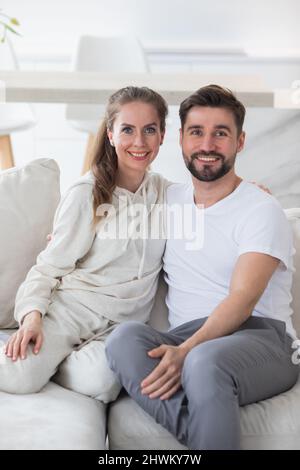Portrait of smiling young man and woman spouse renters sit relax on comfortable couch in new apartment or house. Happy Caucasian couple rest on sofa i Stock Photo