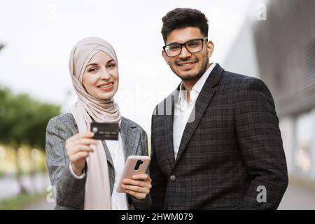 Smiling satisfied multiracial business couple buying online with credit card standing outside airport terminal. Cheerful young colleagues enjoying online shopping of tickets for business trip. Stock Photo