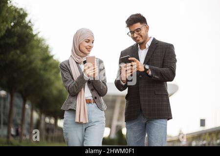 Business people on coffee break using phones outdoor. Arab Muslim male and female colleagues walking on the street and using social networks on smartphones, sharing good news Stock Photo