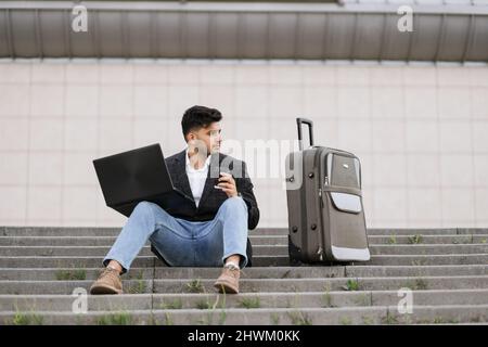 Young arab businessman working on his laptop computer, while sitting on the stairs in the street, with his suitcase, waiting for a business trip and drinking coffee. Stock Photo