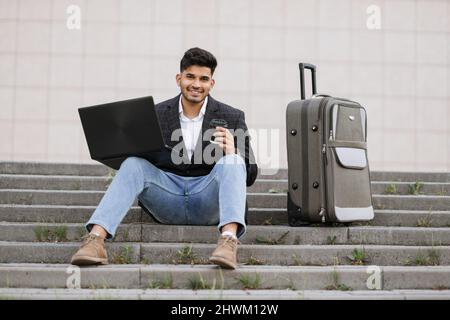 Young handsome bearded Asian Indian man in smart casual clothes with wireless laptop sitting on stairs outdoor near his suitcase, thoughtfully working with coffee and smiling at camera. Stock Photo