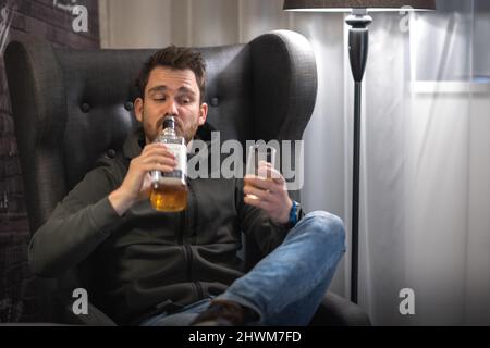 Young drunk is drinking whiskey from bottle, not using a glass. Alcohol abuse, young male heavy drinking whisky in a room Stock Photo