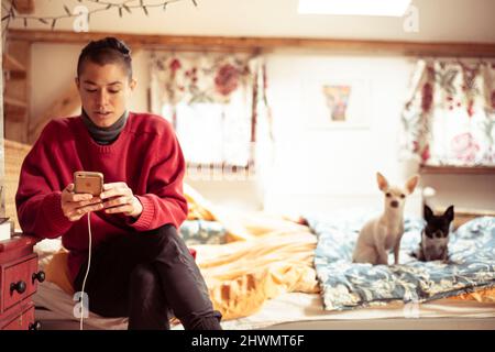 Alternative androgynous person on mobile device in room with two dogs Stock Photo