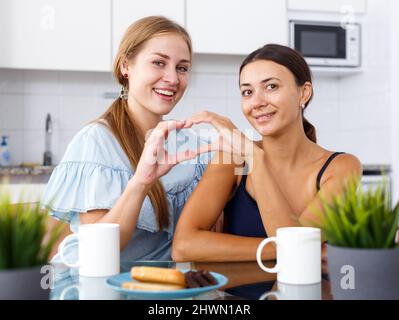Young smiling emotional girlfriends showing the heart with hands and drinking tea Stock Photo