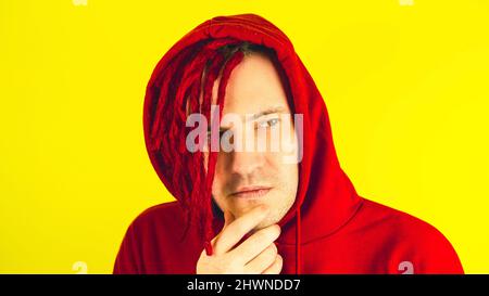 Young man with red dreadlocks in hood with thoughtful look. Stylish male scratching chin and looking away Stock Photo