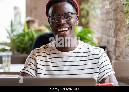 Laughing African American hipster man using laptop, talking online in video chat with friends in cafe Stock Photo