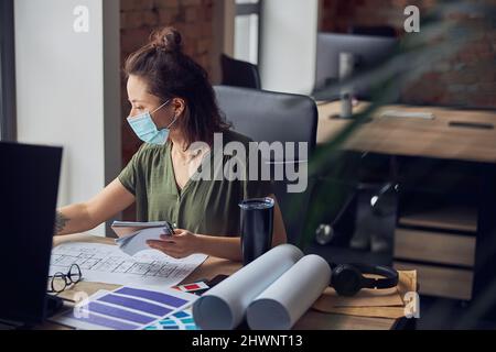 Woman interior designer wearing protective mask making notes in her notebook while working on new project, sitting at the table in her office Stock Photo