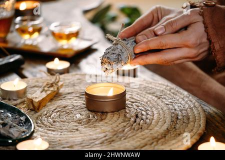 Woman hands burning white sage, before ritual on the table with candles and green plants. Smoke of smudging treats pain and stress, clear negative Stock Photo