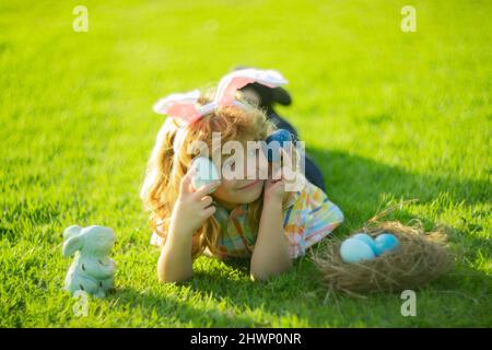 Cute child bunny wear rabbit ears laying on grass in park, Happy Easter day. Bunny boy, funny outdoor portrait. Hunting eggs and spring holidays with Stock Photo
