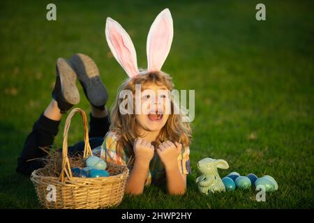 Child bunny boy with rabbit bunny ears. Children hunting easter eggs. Kid boy lying on the grass and finding easter eggs. Child with easter eggs and Stock Photo