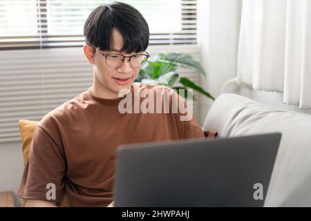 Technology Concept The man that is putting on glasses leaning his back on the dark yellow while focusing on the laptop’s screen. Stock Photo