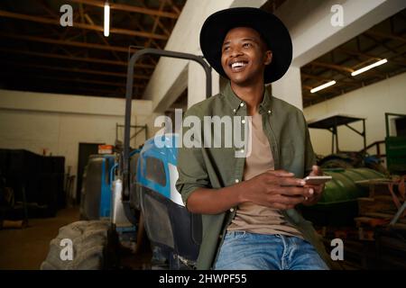 smiling mixed race male farmer sitting on tractor texting on cellular device Stock Photo
