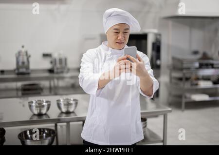 Handsome asian chef cook in white uniform texting on phone, communicating online during a break in the restaurant kitchen. Leisure time during job at restaurant Stock Photo