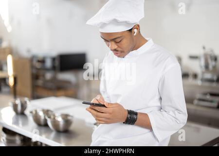 Handsome chef in uniform standing with phone in professional restaurant kitchen. Latin guy using phone on work at restaurant Stock Photo