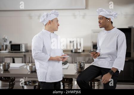 Two multiracial chef cooks talking during a break at the professional kitchen. Asian and Latin chefs in white uniform at work in restaurant. Leisure time and communication of two workers  Stock Photo