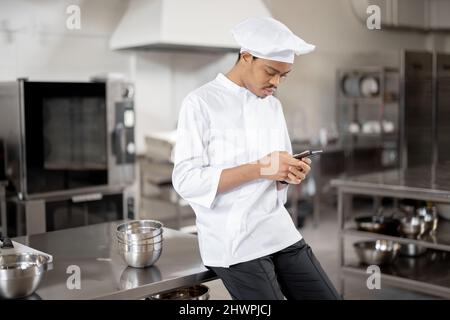 Handsome chef in uniform standing with phone in professional restaurant kitchen. Latin guy using phone on work at restaurant Stock Photo