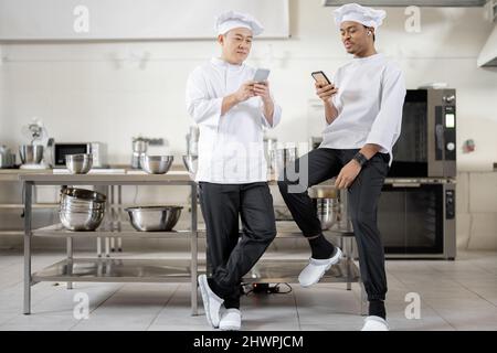 Two multiracial chef cooks standing with smart phones during a break at the restaurant kitchen. Asian and Latin chefs in white uniform during a break at work Stock Photo