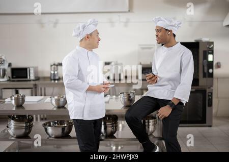 Two multiracial chef cooks talking during a break at the professional kitchen. Asian and Latin chefs in white uniform at work in restaurant. Leisure time and communication of two workers  Stock Photo