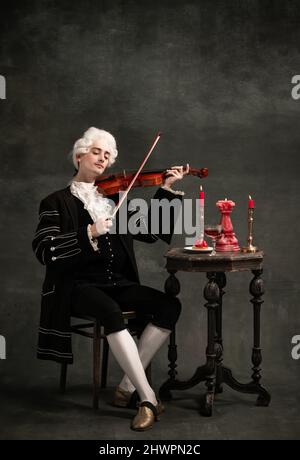 Portrait of young man wearing wig and vintage medieval outfit like famous composer playing violin isolated on dark green vintage background. Stock Photo