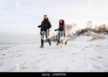 Young couple running with dog on snow at sunset Stock Photo