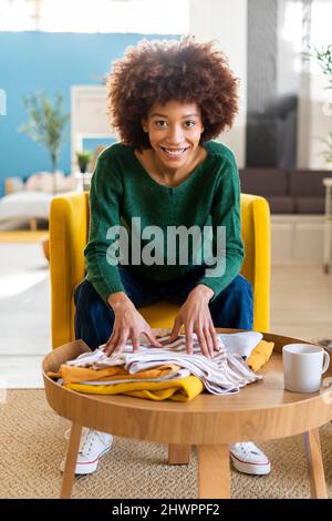 Smiling young Afro woman sitting with folded clothes in living room Stock Photo