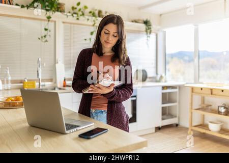Young working woman taking notes in notebook from laptop in kitchen at home Stock Photo