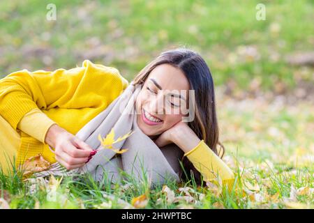 Happy young woman with leaf lying on grass in autumn park Stock Photo
