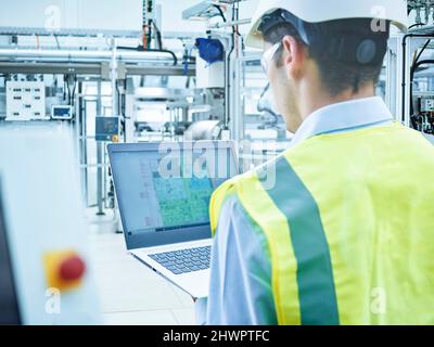 Young engineer in protective workwear using laptop at factory Stock Photo