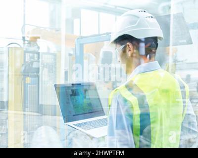Young engineer in protective workwear using laptop seen through glass wall at factory Stock Photo