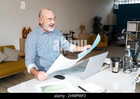 Smiling senior freelancer reading documents sitting in home office Stock Photo