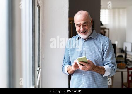 Smiling senior businessman using smart phone by window at home Stock Photo