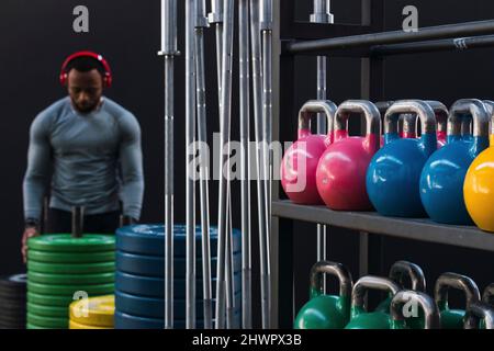 Colorful kettlebells and barbells stacked on rack with athlete in background in gym Stock Photo