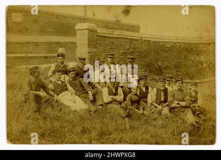 Original sepia-toned Victorian era cabinet card relaxed outdoor portrait of large group of working class men and boys (apprenticed maybe) sitting near an ancient harbour wall near a slipway. They are all wearing peaked mariners caps, and some are wearing hand knitted fishermen's sweaters, another an apron, and are probably in the fishing industry - a coastal seafaring fishing community in the U.K. Dated June 1892. Stock Photo
