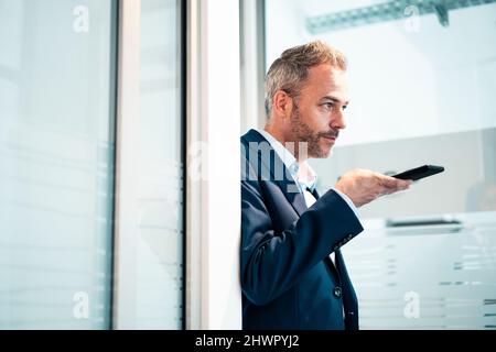 Businessman talking through smart phone speaker leaning on doorway at office Stock Photo