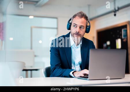 Thoughtful businessman wearing wireless headphones using laptop at desk in workplace Stock Photo