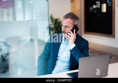 Worried businessman sitting with laptop at desk talking on smart phone Stock Photo