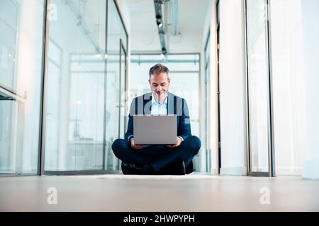 Businessman sitting cross-legged using laptop in office corridor Stock Photo