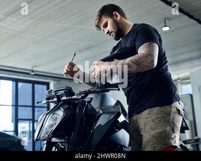 Tattooed mechanic with tool working on motorcycle in workshop Stock Photo