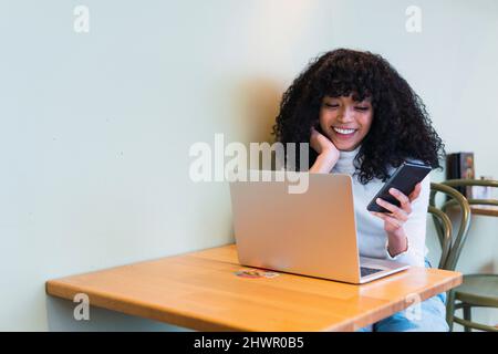 Happy young curly haired woman using smart phone sitting with laptop at table in cafe Stock Photo