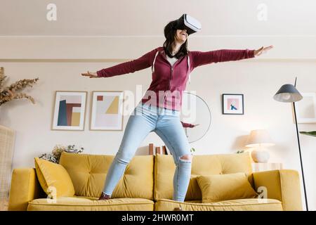 Happy woman wearing virtual reality headset standing with arms outstretched on sofa Stock Photo