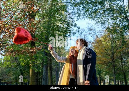Smiling couple holding red heart shape balloon at park Stock Photo