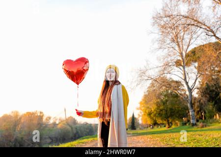 Smiling redhead woman holding red heart shape balloon in autumn park Stock Photo