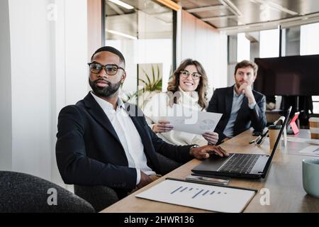 Young businessman wearing eyeglasses sitting by colleagues at conference table in board room Stock Photo