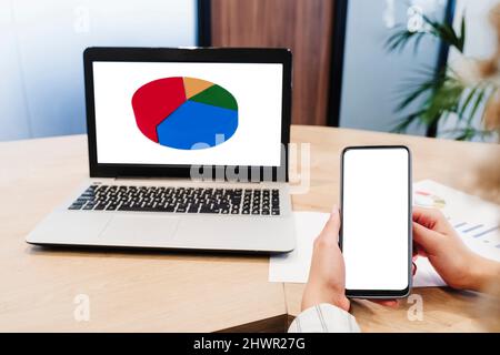 Hands of businesswoman showing blank screen of smart phone near laptop with pie chart at office Stock Photo