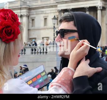 Stand with Ukraine rally in Trafalgar Square, London 2022. Lady paints Ukrainian flag on mans face to show solidarity with Ukraine. Stock Photo