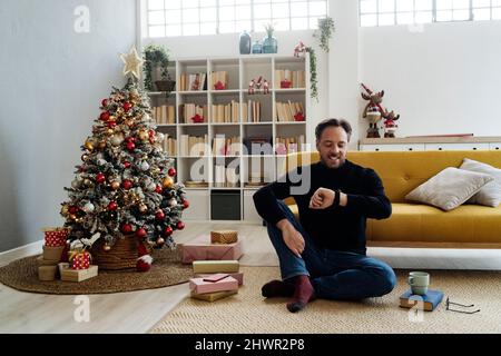 Happy man checking smart watch sitting by Christmas tree at home Stock Photo