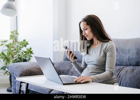 Pregnant woman with smart phone working on laptop at home office Stock Photo