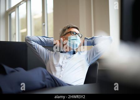 Businessman with hands behind head wearing protective face mask at office Stock Photo