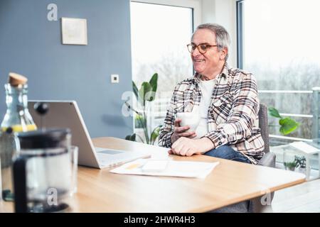 Smiling senior man holding coffee cup watching videos on laptop at home Stock Photo